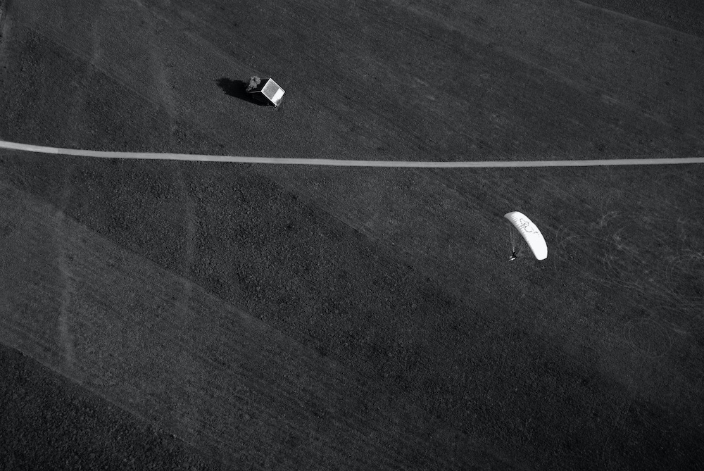 Paraglider captured from above flying over a field with a mountain hut